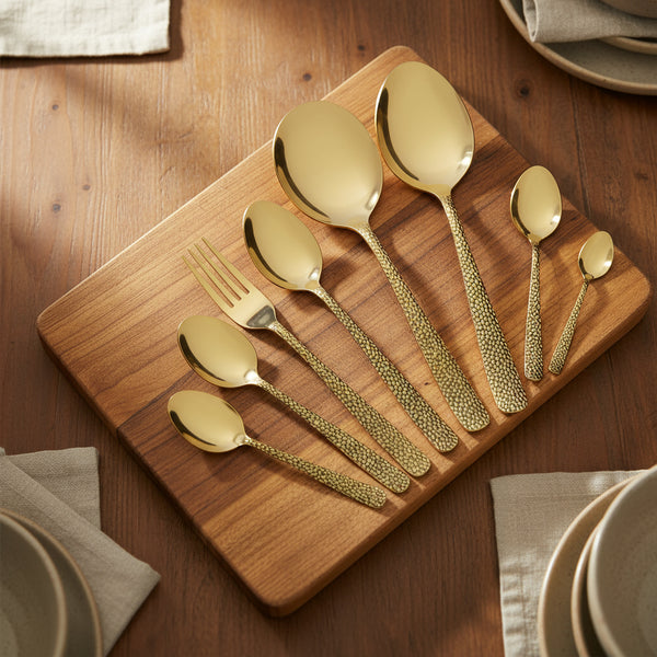 Set of gold flatware on a wooden cutting board with plates and napkins in the background.