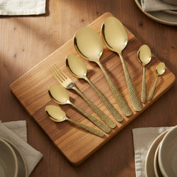 Set of gold flatware on a wooden cutting board with plates and napkins in the background.